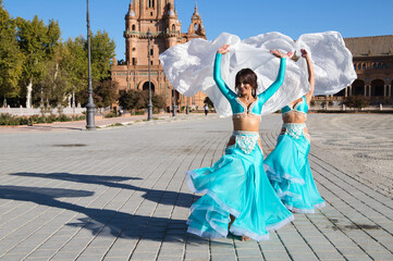 Two young and beautiful belly dancers dancing in a square. They are dressed in light blue with...