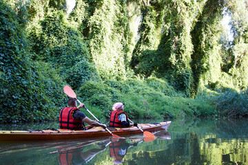 Couple man and woman paddle a orange kayak in wilderness areas at Danube river among green trees, wild grapes and cane at spring high water on Danube biosphere reserve