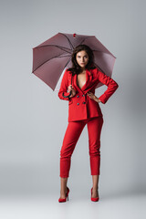 full length of brunette woman in red suit standing with hand on hip under umbrella on grey.
