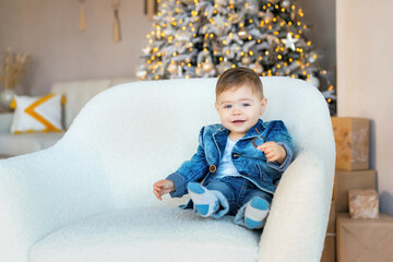 Fototapeta premium Happy laughing one-year-old child wearing stylish blue jacket and jeans sitting in a chair near the Christmas tree. Room with Christmas decorations. White and gold decor. Happy New year