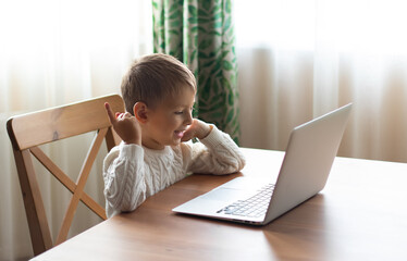 Little boy in a white sweater is sitting at a laptop and speaking by video call.