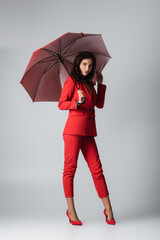 full length of young woman in red suit standing under umbrella on grey.
