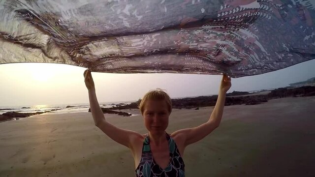 Woman walks on the beach at sunset. Pretty middle-aged female tourist dancing on seashore, pareo develops in her hands.