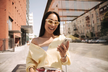 Closeup happy girl looks at phone screen and smiles while standing outdoors. She is thrilled to...