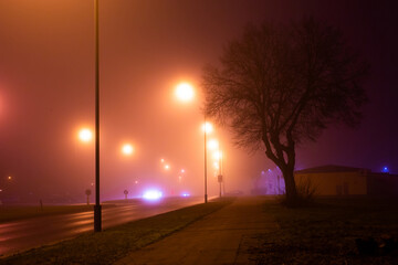 Foggy mysterious misty city road with mild pink yellow and orange street lights and silhouette of the tree. Nobody on the street. Good as background or wallpaper