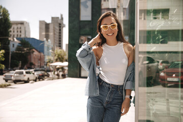 Summer sunny image of young stylish hipster woman standing near wall of panoramic building. Brunette in cute light denim clothes is resting and enjoying weekend.