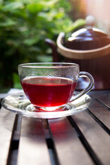 Glass cup of hot red hibiscus tea and and old retro teapot on a wooden tray at terrace. Close up