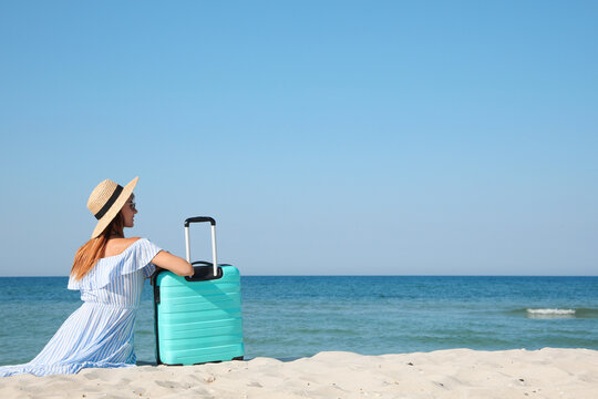 Woman with suitcase sitting on sandy beach near sea, back view