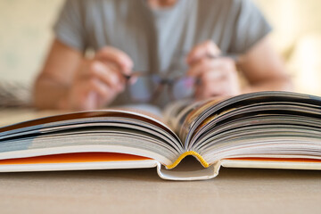 Close-up view of open book lying on table and male hands holding eyeglasses in blurred background