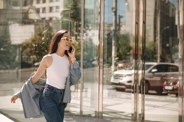 Fototapeta premium Asian woman walks in flying gait along panoramic windows of office. Brunette in sunglasses talking on phone with relatives. Dressed in white blouse, jeans and shirt.