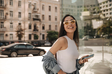 Stunning asian woman poses for photo standing half sideways with dark hair on street of sunny city. Girl in white top, blue shirt on her shoulders with phone in her hands.