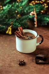 A mug of hot chocolate or cocoa with cinnamon sticks and pieces of chocolate on the background of fir branches.Christmas and winter holidays.Glass of winter drink on rusty background