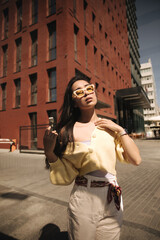 Beautiful sophisticated asian lady is walking under rays of sun against backdrop of red house. Closed-eyed brunette with glasses is wearing white top, yellow jacket on top and long shorts with belt.