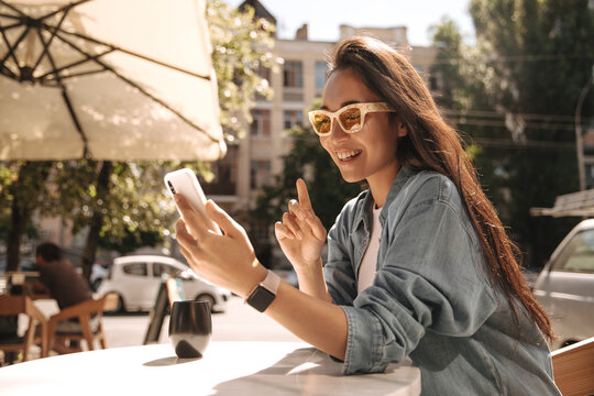 Cute young asian brunette in glasses looking at smartphone sitting in cafeteria on street. Happy girl student of university using mobile phone communicates by video communication with friends.