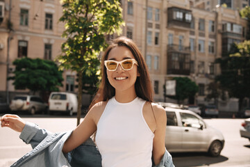 Pretty light-skinned young asian brunette in sunglasses posing for photo. Smiling girl in top and blue shirt on top is walking streets of city. Good mood, fashion trends