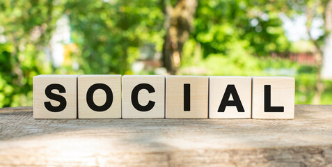 Six wooden blocks lie on a wooden table against the backdrop of a summer garden and create the word SOCIAL.