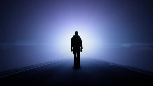 Empty road with a standing man and a traffic flashing light. Silhouette of a male pedestrian on a dangerous foggy asphalt road with low visibility.