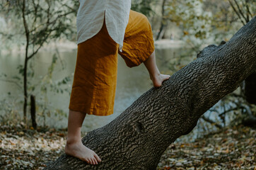 Person in saffron linen pants balancing warrior stance on a fallen tree trunk, barefoot connection...