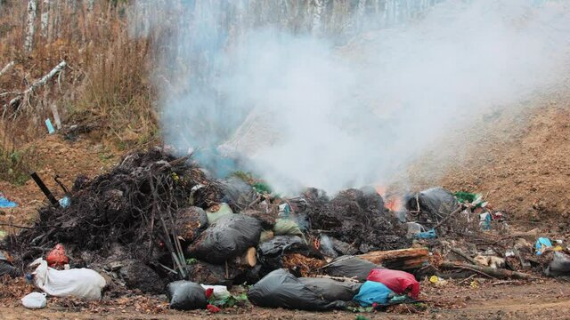 Old garbage is lying on the ground and burning. Acrid and fetid smoke rises above this pile. Air and soil pollution at the city landfill.