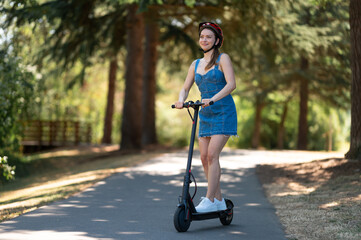 A nice girl in a protective helmet and denim dress rides an electric scooter in the park. Clear sunny day, beautiful nature. Eco-friendly transport, outdoor activities, sports. © Anton