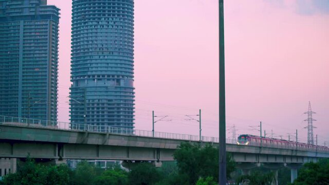 Dusk shot of metro train on elevated bridge tracks moving toward camera with high rise multistory skyscrapers in the background with pink dawn clouds in Noida gurgaon delhi