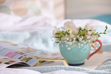 Beautiful bright flowers in cup, open magazine and tray on bed. Space for text