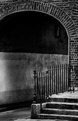 Arched brick entrance to narrow lane showing wall beside railing and  steps. Monochrome black and...