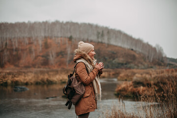 Naklejka premium A young girl tourist in brown leggings, a brown jacket, a beige hat and a brown backpack in the fall walks by the river, overlooking the forest, drinks tea from a mug