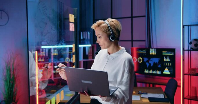 Beautiful confident motivated experienced blond businesswoman in headset communicating with remote interlocutor while working on glass wall with datas from computer in office