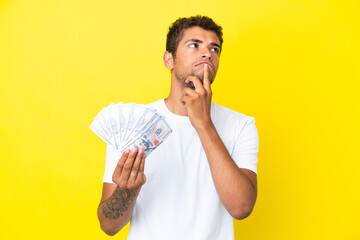 Young brazilian man taking a lot of money isolated on yellow background having doubts while looking up