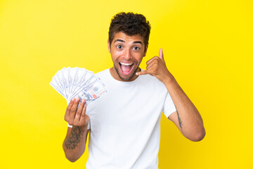 Young brazilian man taking a lot of money isolated on yellow background making phone gesture. Call...