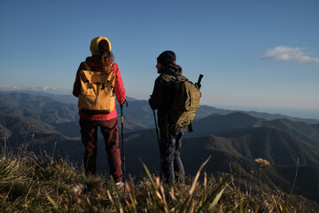 Fototapeta na wymiar Travel in mountains with loved one and enjoy beautiful panoramic view. Man and woman travelers in red jacket with yellow backpack are standing with their backs turned and looking at mountains.