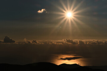 Sun illuminates sea with its bright orange light and you can see silhouette of ship on horizon. Beautiful seascape with mountains and sunset rays in distance.