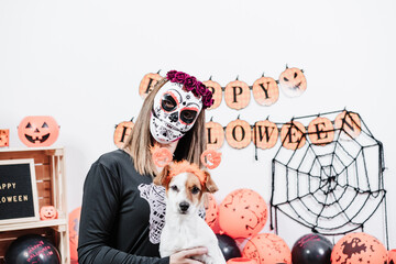 jack russell dog with funny ghost costume and woman wearing mexican face mask during halloween celebration. woman wearing skeleton costume and red roses diadem on head. Halloween party concept