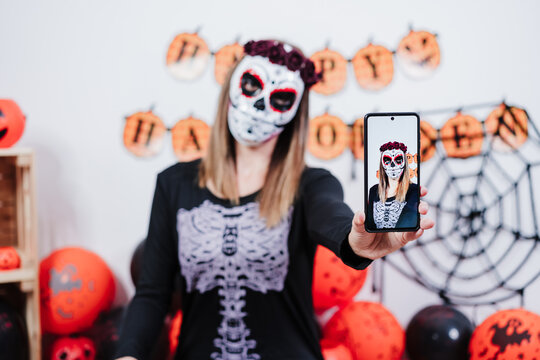 woman wearing mexican face mask during halloween celebration. taking selfie with mobile phone. woman wearing skeleton costume and red roses diadem on head. Halloween party concept