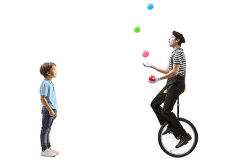Full length profile shot of a little boy looking at a mime juggling with balls and riding a unicycle - obrazy, fototapety, plakaty