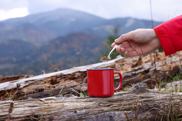 Woman pouring instant coffee into mug in mountains, closeup. Space for text