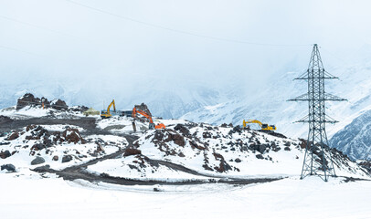 Three excavators are working in the snowy mountains next to the power line. Construction work, business in difficult conditions. Preparation of a site for the construction of a ski resort.