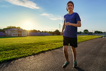 concept of sports and health - teen boy runs along the stadium track, a soccer field with green...