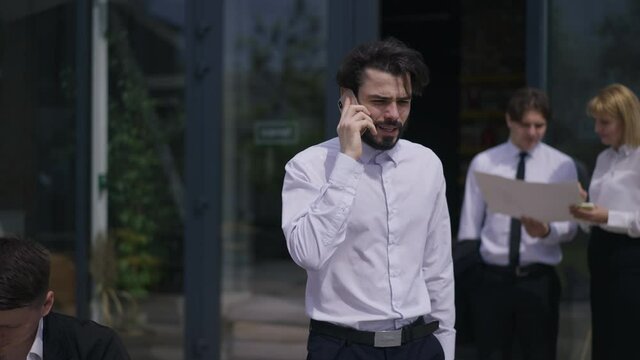 Portrait of confident bearded man persuading partners talking on phone walking in slow motion passing colleagues. Handsome Caucasian employee discussing partnership on office terrace outdoors