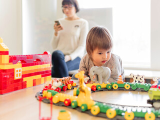 Toddler plays with colorful toy blocks while his mother or babysitter texting in smartphone. Little boy stares on toy constructor. Interior of kindergarten or nursery.