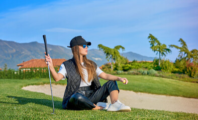 Smiling woman golf player putting successfully ball on green, ball dropping into cup, blue sky in...