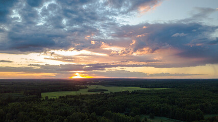 Aerial landscape of sundown over the forest and green fields. Bright orange sky during a sunset with dark clouds. 