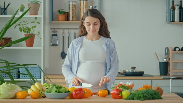 Happy Pregnant Woman Enjoying Preparing a Salad, Cutting Organic Vegetables in Home Kitchen With Modern Interior. Healthy Eating During Pregnancy. A Pregnant Woman Prepares Food at Home.