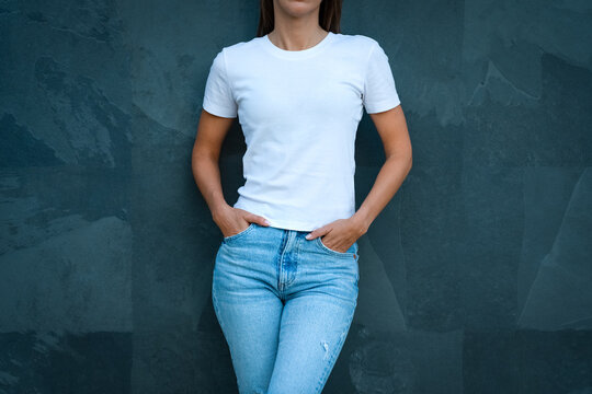 Young woman dressed in blank white t-shirt and blue jeans posing against the grey wall. Mockup for design print or t-shirt print store