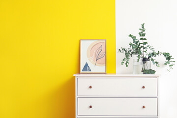 Vase with eucalyptus branches and picture on chest of drawers near color wall