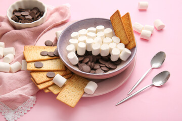 Plate with crackers, marshmallows and chocolate on pink background