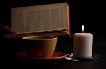 A candle lit with a flame, with a cup of tea with smoke, and a reading book in the background. (focus on candle).