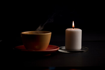 Fototapeta na wymiar candle lit with flame, with cup of tea with smoke, black background. (focus on candle).