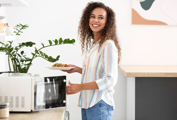 Young African-American woman putting plate with food into microwave oven in kitchen - obrazy, fototapety, plakaty